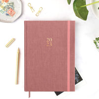 Picture of the 2023 Ponderlily planner Sunday start in Rose on desk with pen
